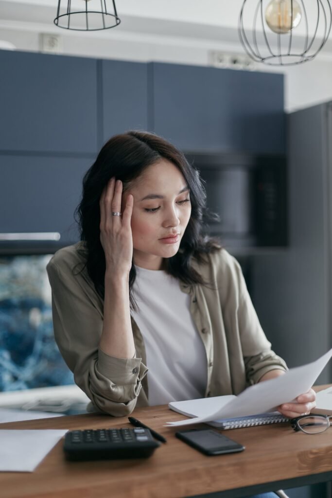 Woman reading paperwork at a kitchen table, resting one hand against her head, with a calculator and phone nearby.