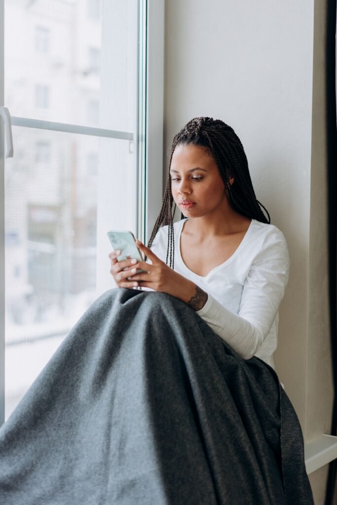 A woman sits beside a window under a gray blanket, looking at her smartphone.
