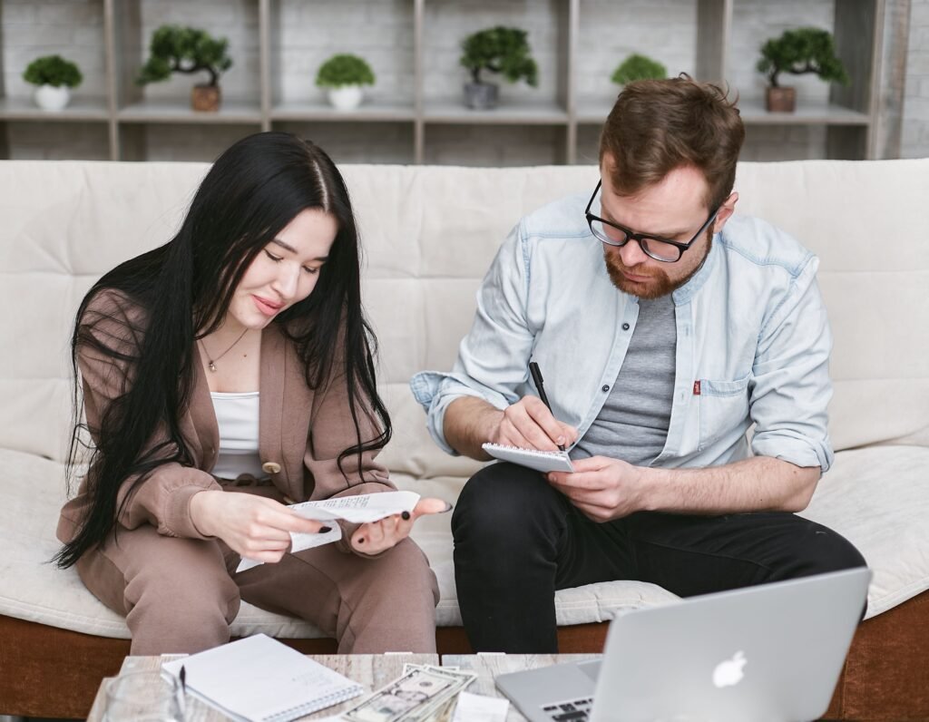 Couple sitting together on a sofa, reviewing bills and writing notes while planning their household budget.