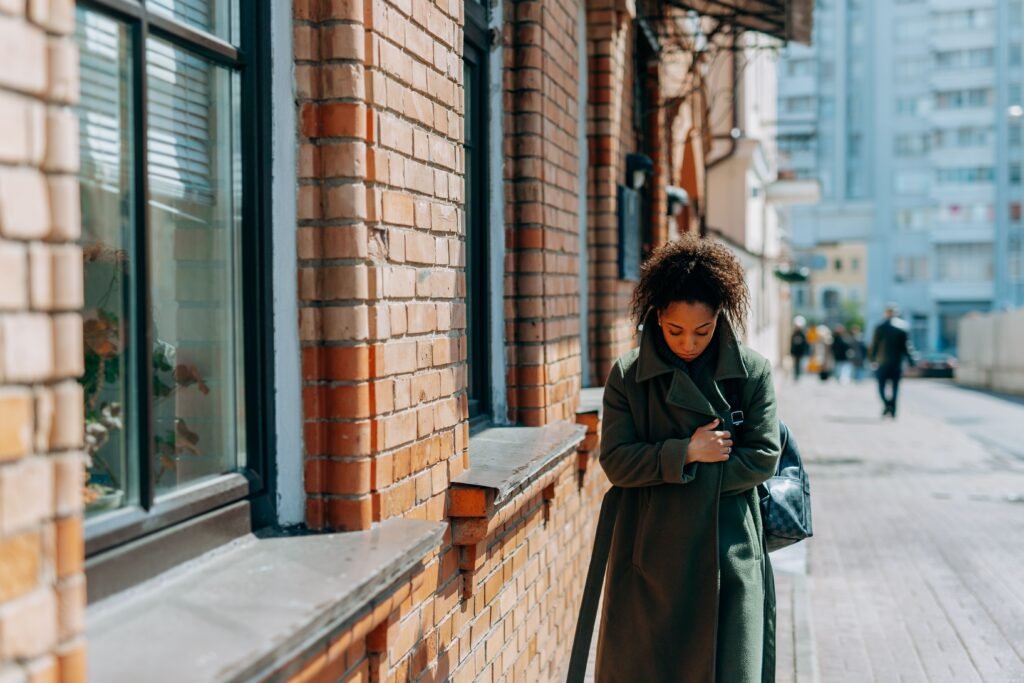 A woman in a green coat walks beside a brick building, looking down.