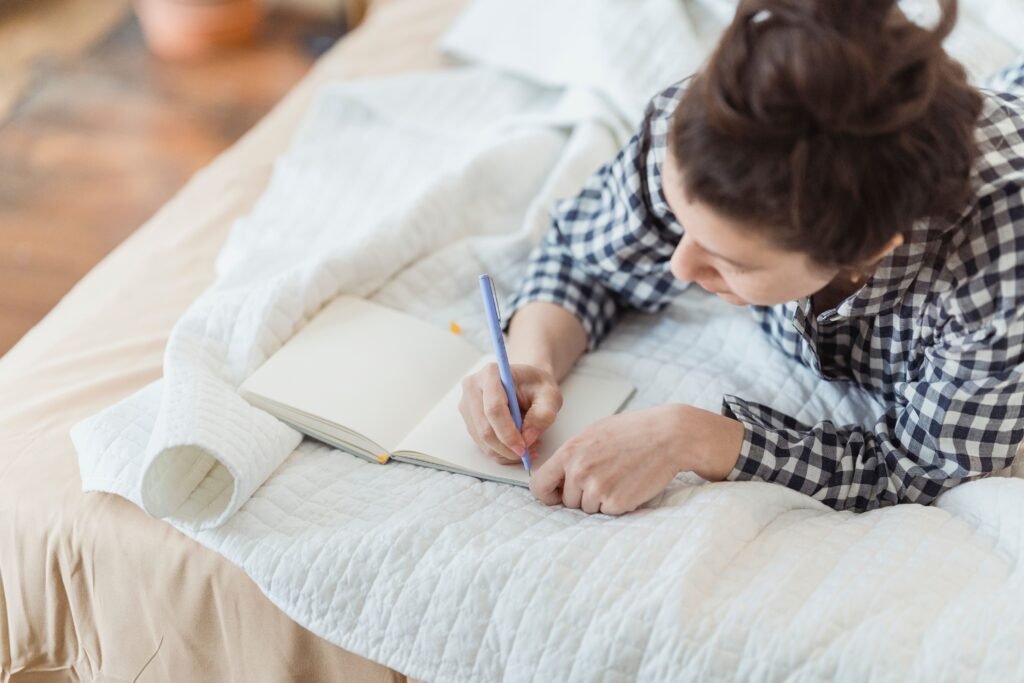 Woman in a checked shirt lying on a bed and writing in an open notebook.