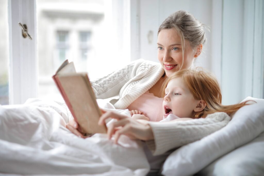 Mother cuddling with her young daughter in bed while reading a book together in a bright, cozy bedroom.