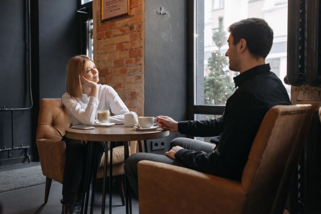 A woman and man talk over coffee at a small café table beside a window.