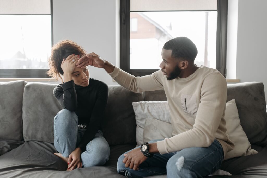 Man gently touching a woman's forehead as they sit together on a sofa, showing care and emotional support.