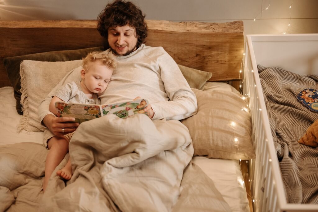 Man reads a picture book in bed while a young child rests against him.