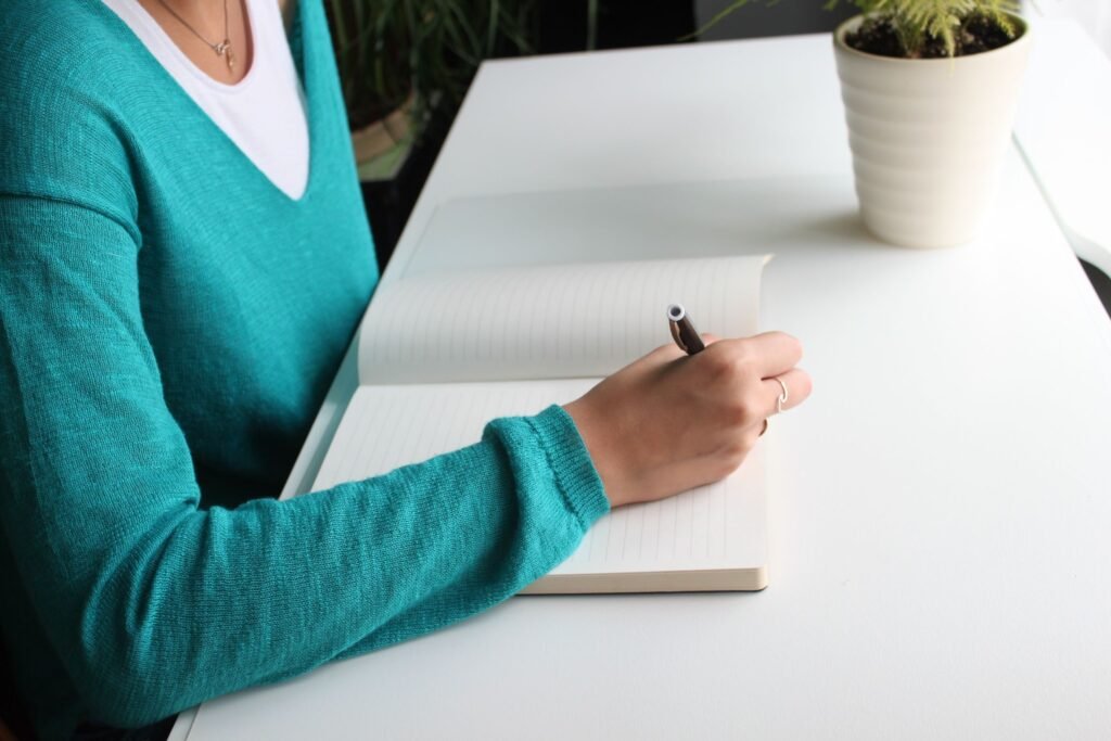 A person in a turquoise sweater writes in an open notebook at a white desk beside a potted plant.