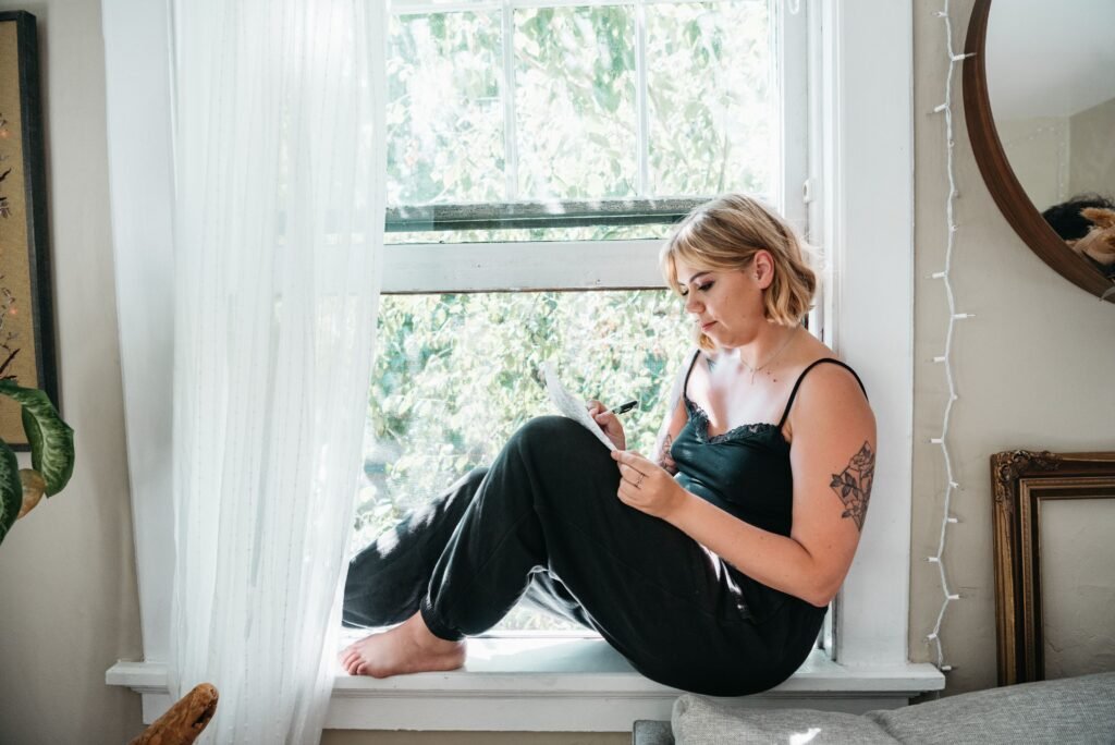 Woman sitting by a bright window, writing notes on a piece of paper while enjoying a quiet moment alone.