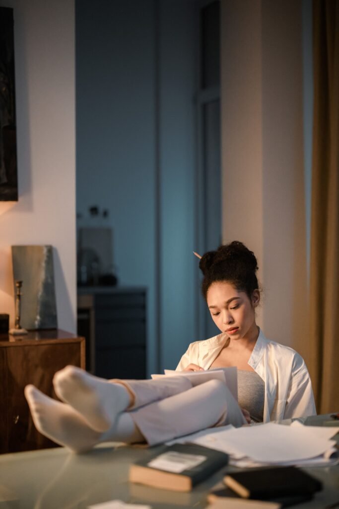 A woman sits with her feet resting on a desk, looking down at papers in a warmly lit room.