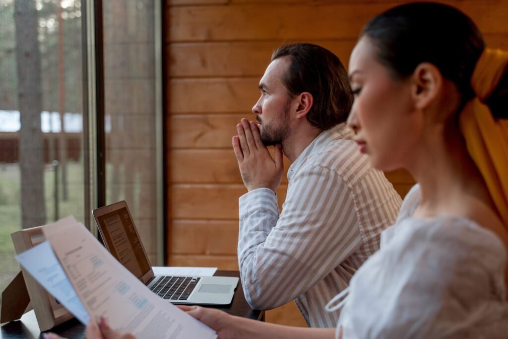 A woman reads papers while a man sits beside a laptop and looks out the window.