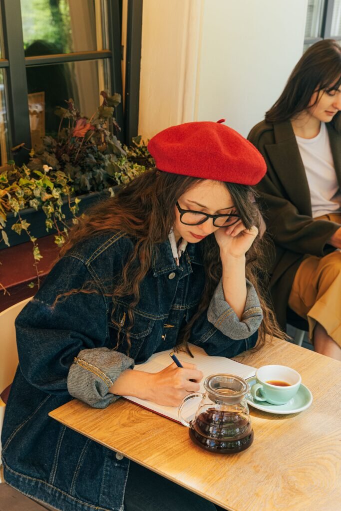 A woman wearing glasses and a red beret writes in a notebook at a café table beside a teapot and cup.