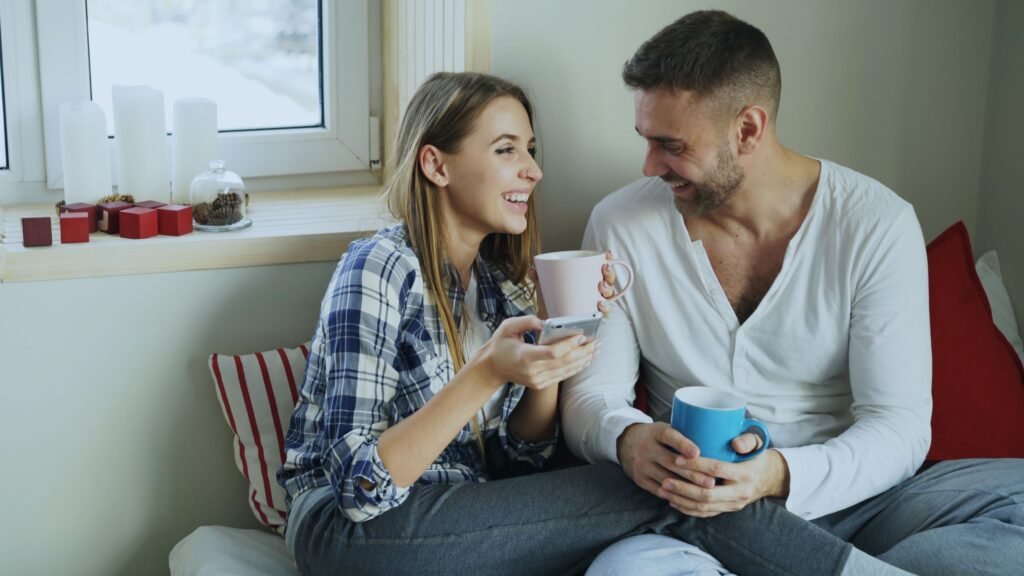 A smiling man and woman sit together holding mugs while the woman holds a smartphone.