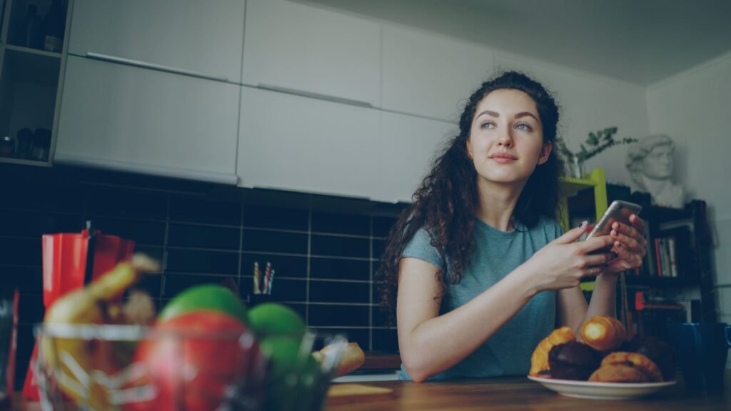 Woman sitting at a kitchen table holding her smartphone and looking thoughtfully into the distance.