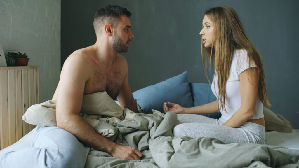 Man and woman sitting facing each other in bed, with the woman gesturing as they talk.