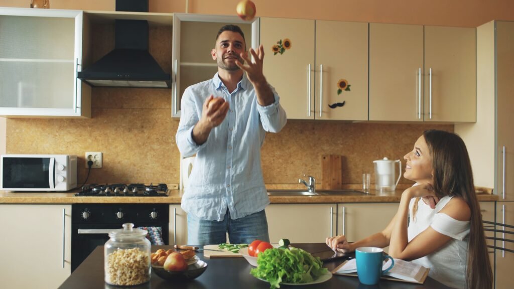 Man playfully tossing an apple in the kitchen while a woman sits at the table smiling and watching him.