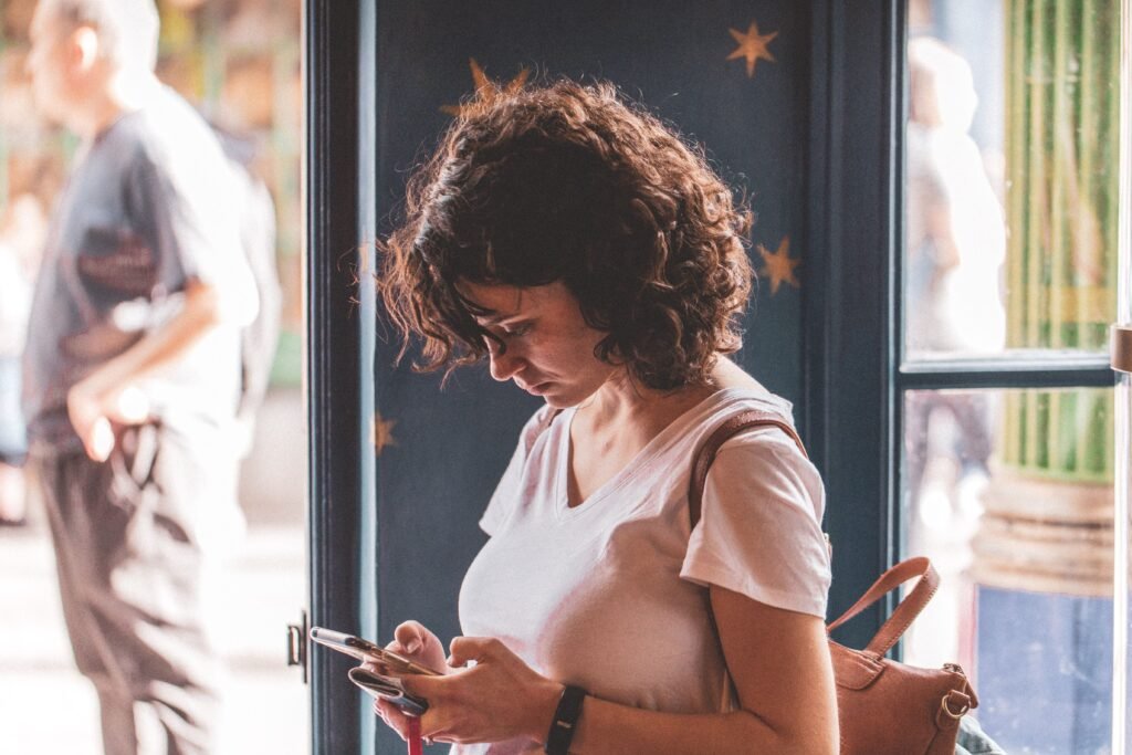 A woman looks down at her phone while standing beside a window.