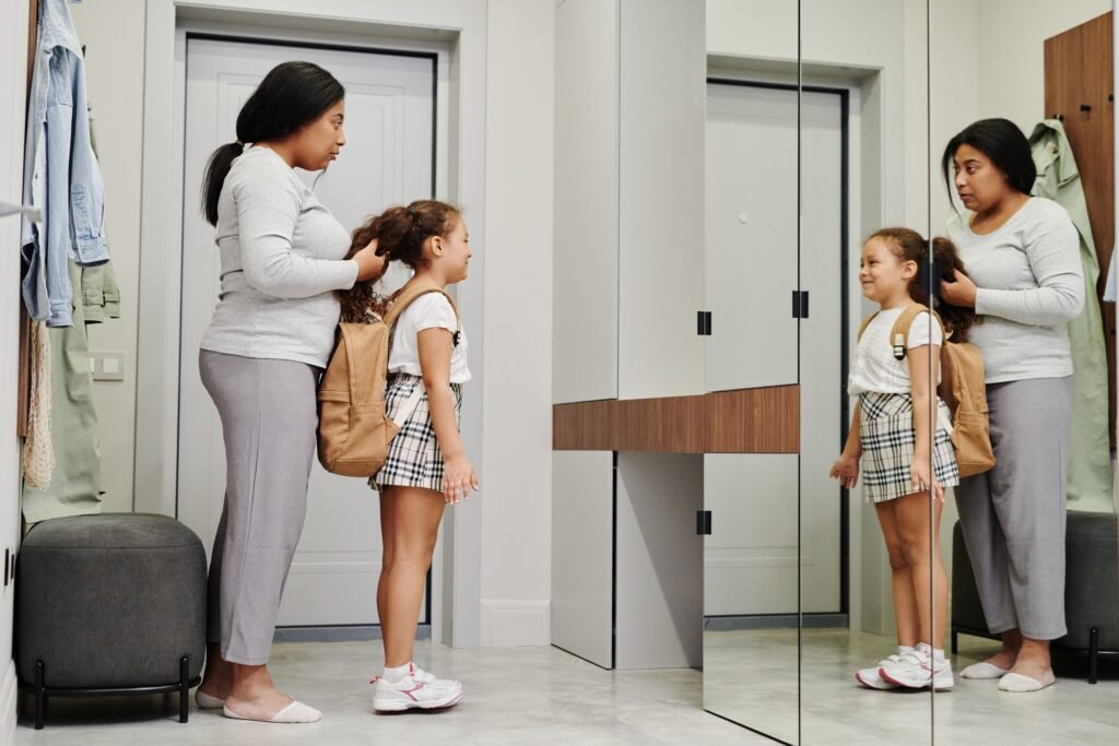 A woman fixes a girl's hair while the girl wears a backpack and looks in a mirror.