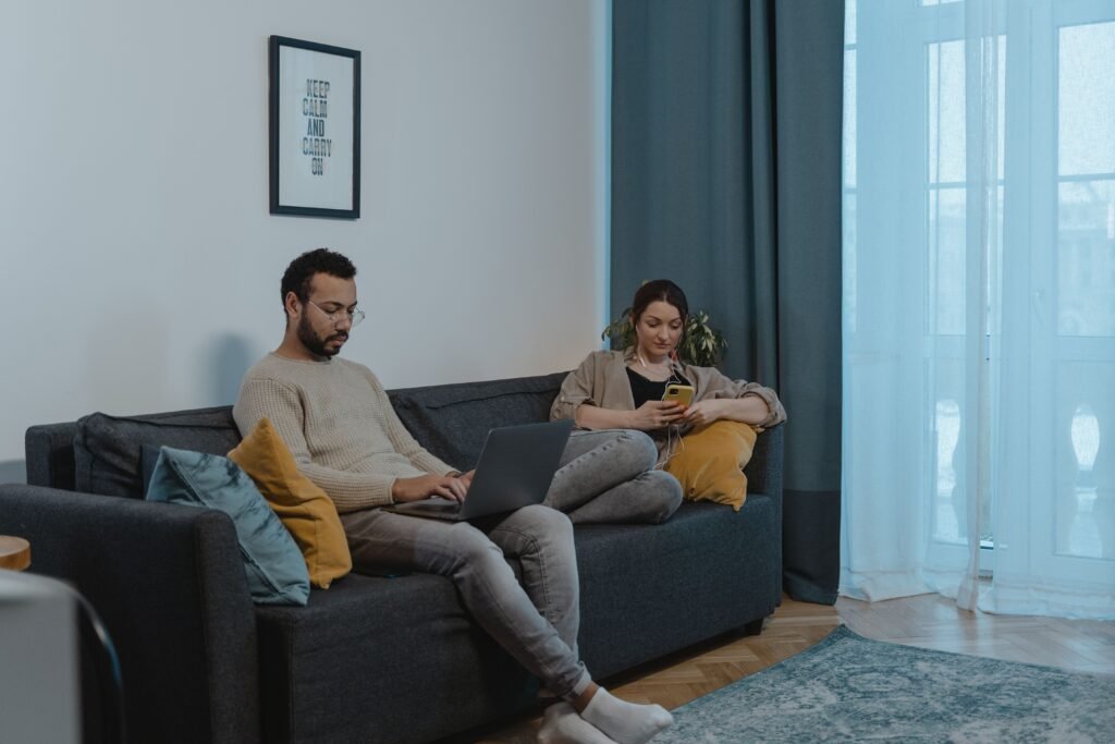 A man uses a laptop while a woman looks at her phone as they sit on a gray sofa.
