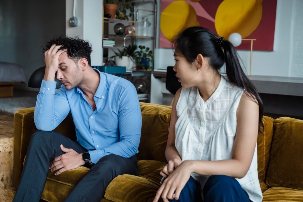 A woman faces a man on a sofa as he looks down with one hand resting on his forehead.