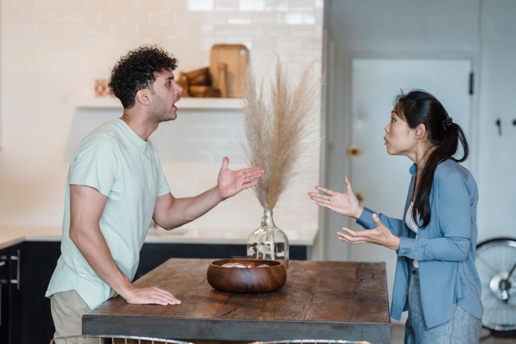 A man and woman face each other across a wooden table, gesturing during a tense conversation.