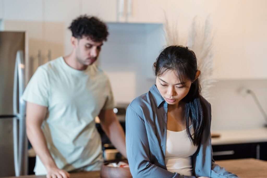 Woman looking down with a concerned expression while a man stands behind her in the kitchen during a tense moment.