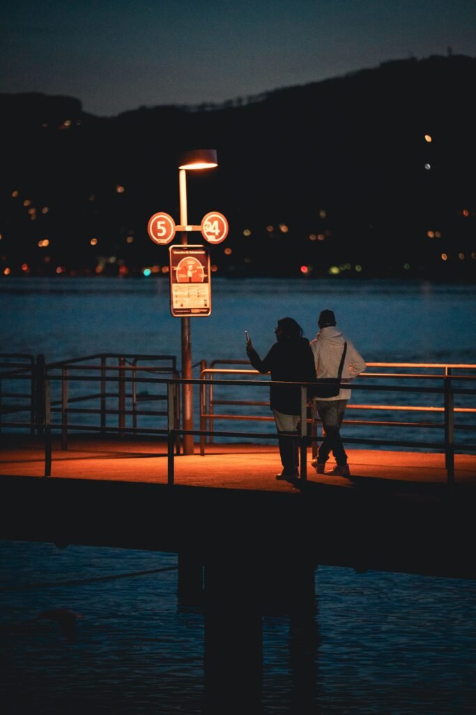 Two people stand beside a waterfront railing at night beneath a glowing lamp.