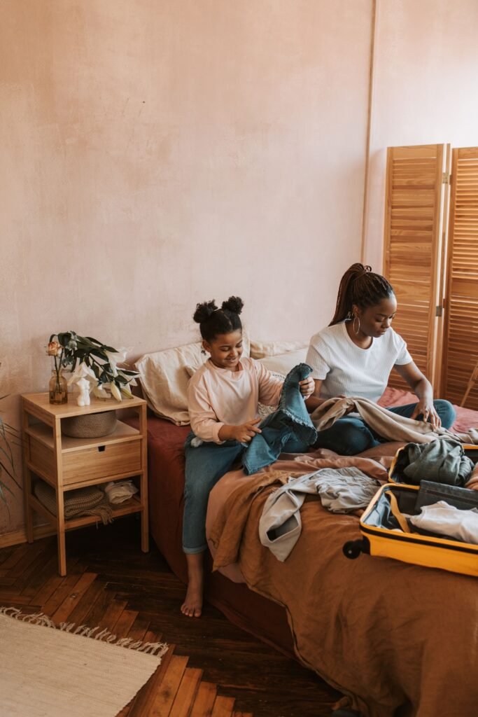 Woman and young girl folding clothes on a bed beside an open yellow suitcase.