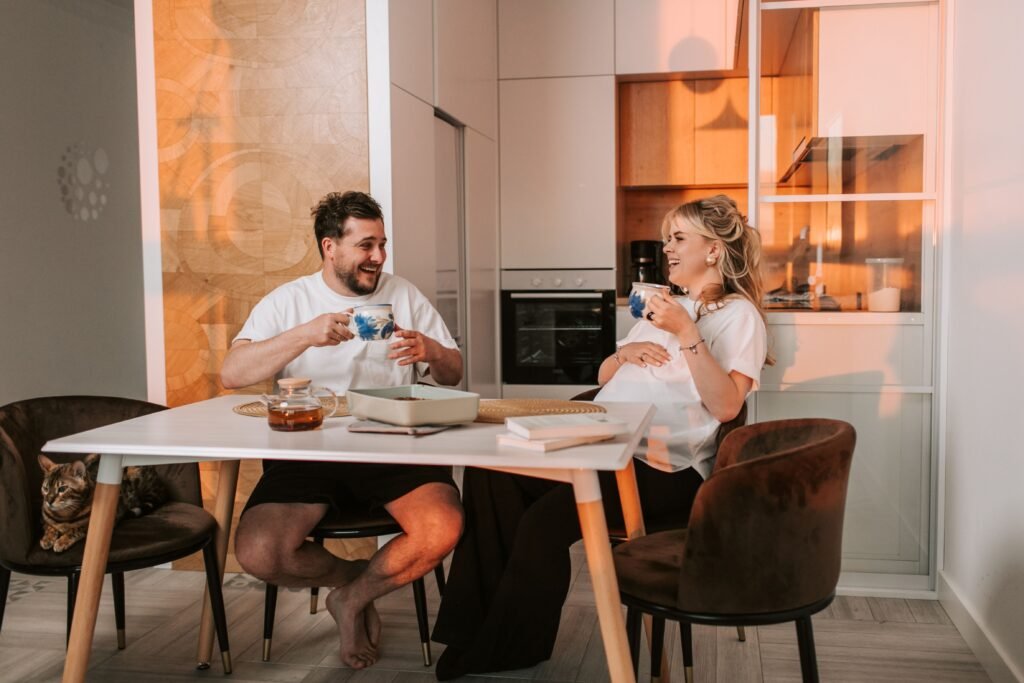 Expectant couple sitting together at a kitchen table, smiling and enjoying a conversation over cups of tea in their cozy home.