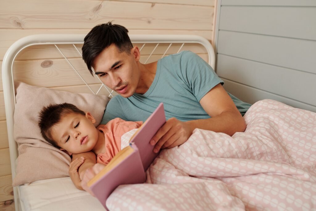 Father reading a bedtime story to his young son while lying together in bed under a cozy pink blanket.