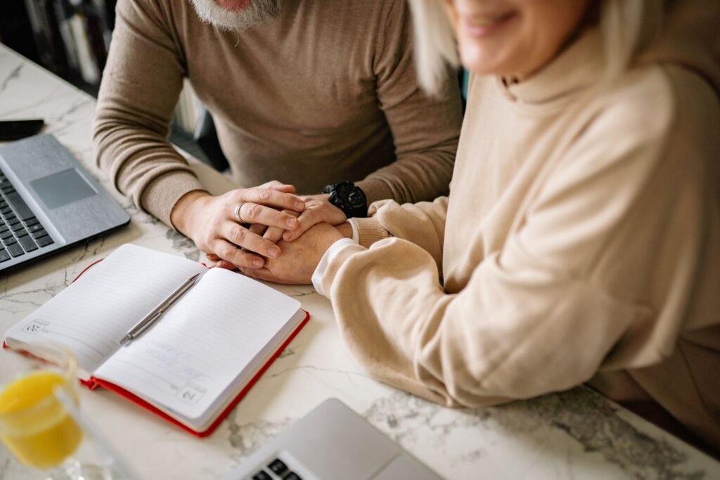 Mature couple sitting together at a table, holding hands beside an open notebook and laptop, sharing a supportive moment.