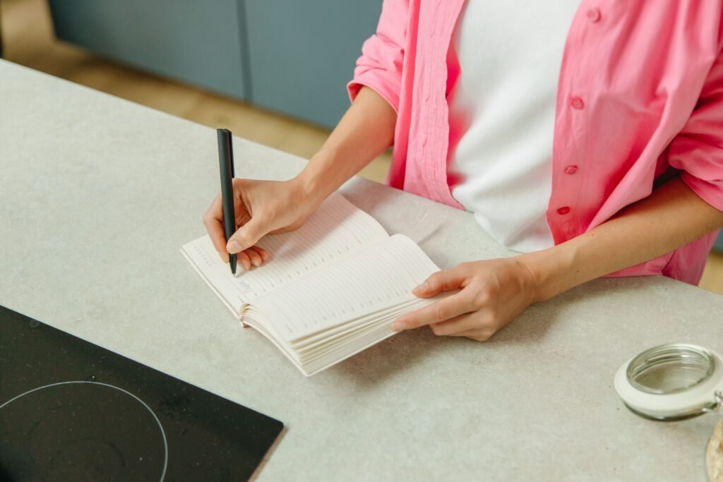Person in a pink shirt writing in an open planner on a kitchen counter.