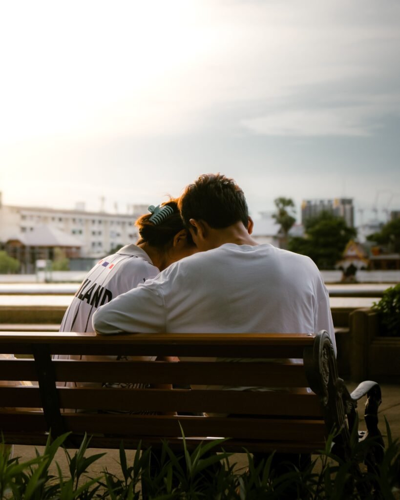 Couple sitting close together on a park bench at sunset, leaning their heads toward each other while enjoying a quiet moment.