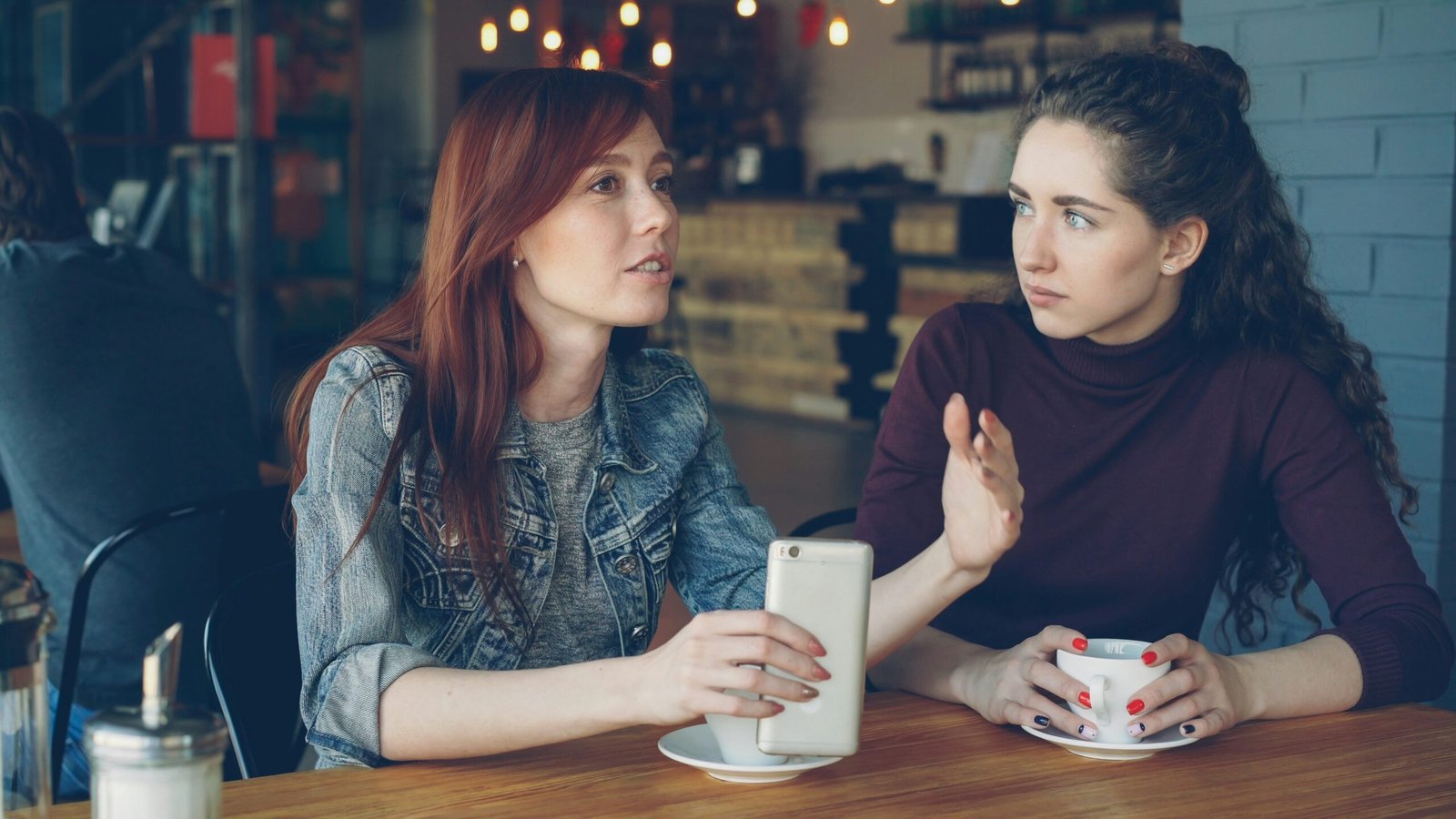 Two women talk across a café table while holding cups.